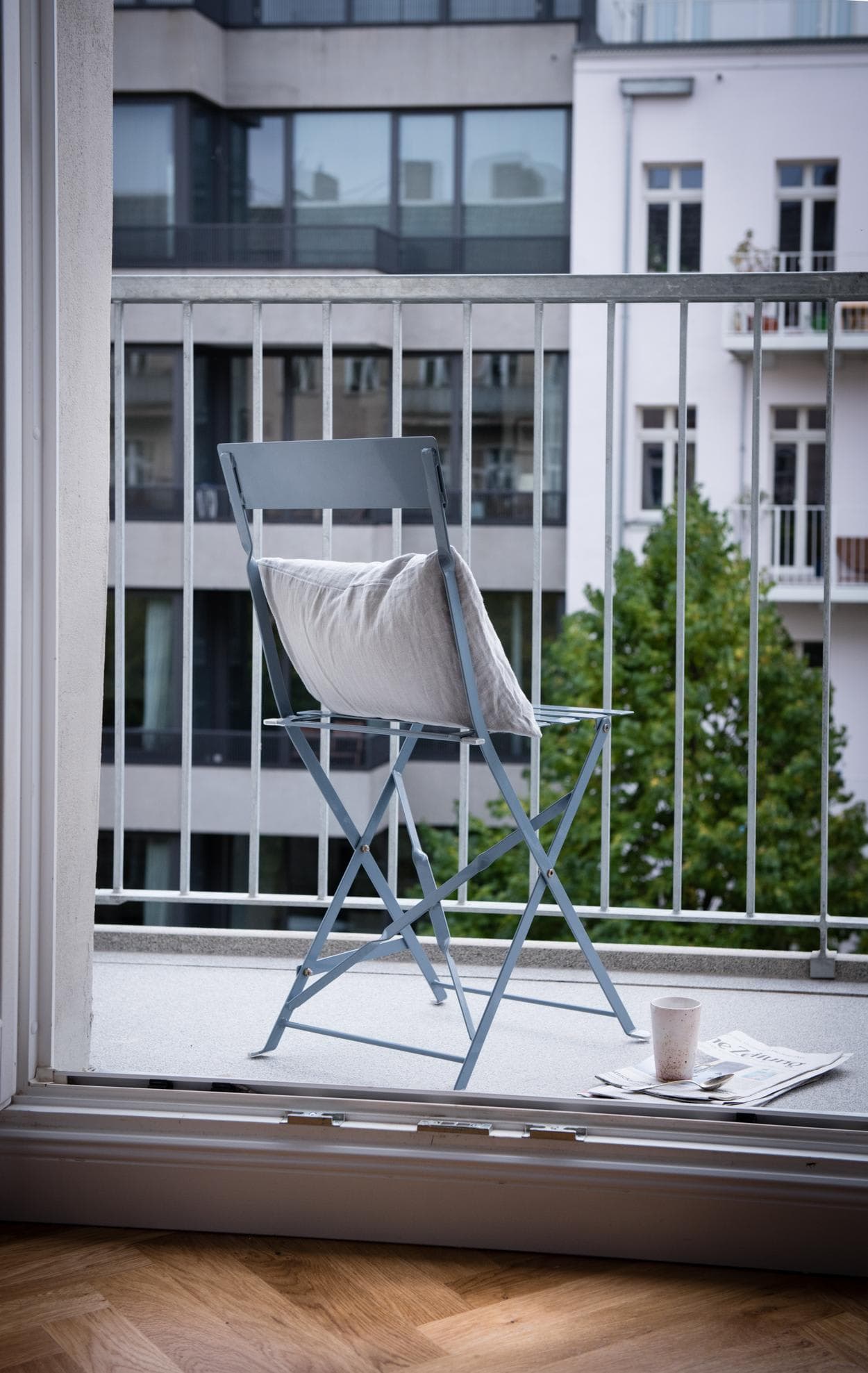 The image features a white lawn chair placed on a wooden deck or balcony, overlooking a cityscape. The chair is situated near the edge of the deck, providing a comfortable spot for relaxation and enjoying the view. The deck is surrounded by a fence, which adds an element of privacy and security to the space.
In the background, there are several buildings visible, including a tall building and a smaller one. The scene appears to be a pleasant outdoor setting, with the chair providing a cozy spot to sit and appreciate the cityscape.