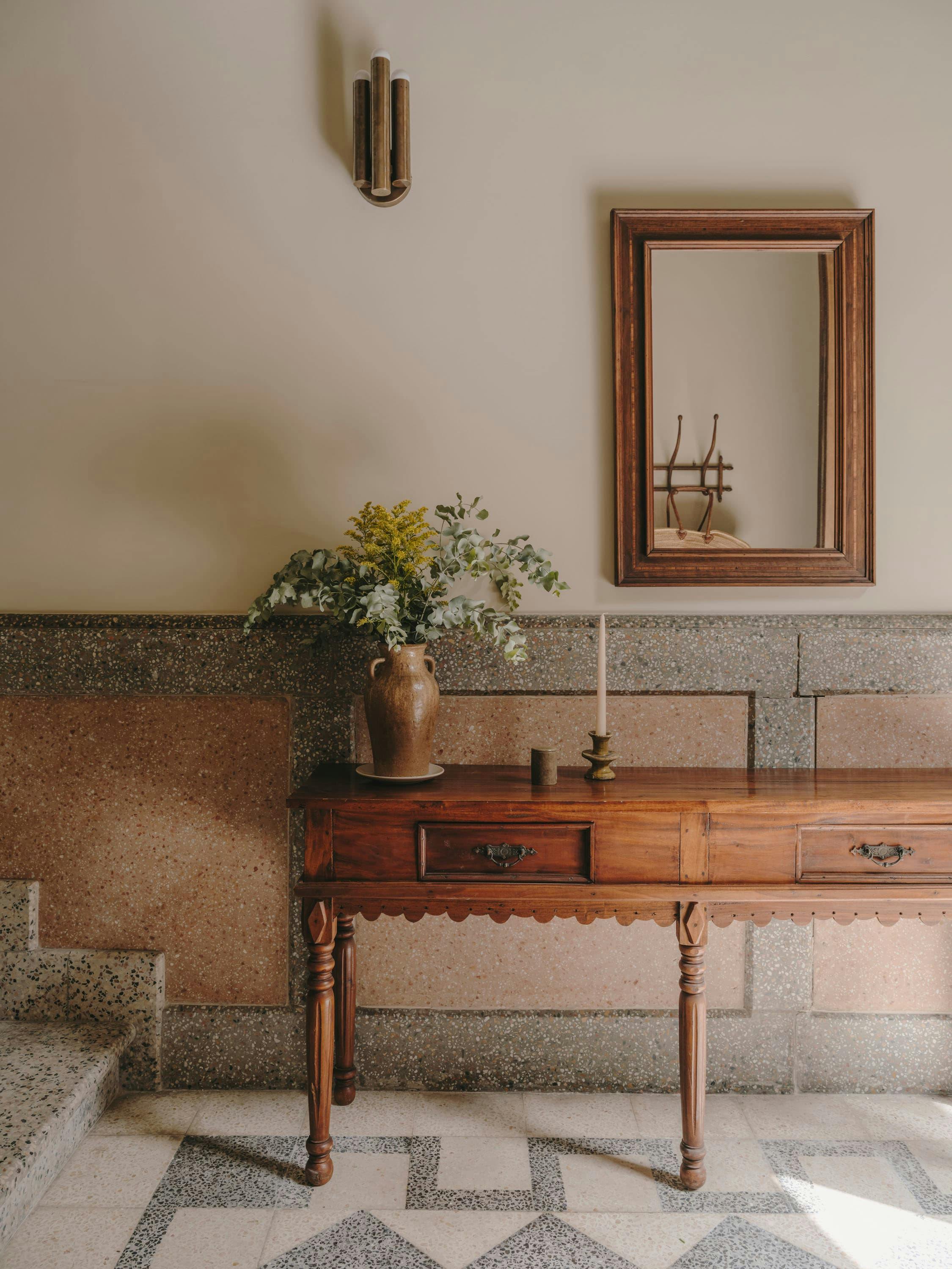 The image features a bathroom with a large mirror above a wooden vanity. The vanity is adorned with a vase of flowers, adding a touch of elegance to the space. The bathroom also has a wooden cabinet and a wooden bench. The room is well-lit, creating a warm and inviting atmosphere.

In addition to the main vanity, there is a smaller vanity located on the left side of the room. The bathroom appears to be clean and well-maintained, with a well-organized layout.
