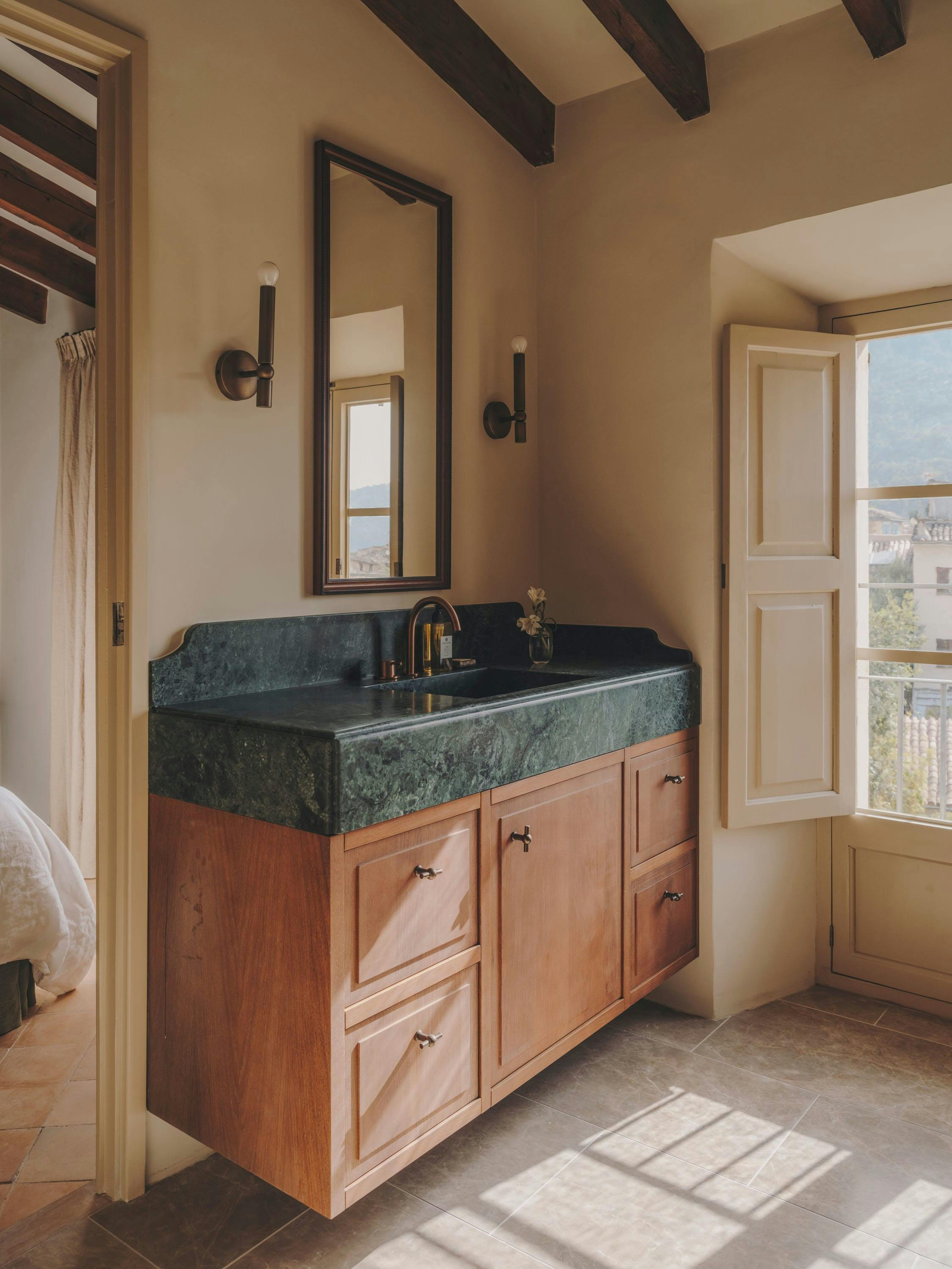 The image features a large, modern bathroom with a double sink vanity. The vanity is made of wood and has a green marble countertop. The sink is placed under a large window, allowing natural light to enter the room. The bathroom also has a large mirror above the sink, adding to the overall aesthetic.

In addition to the sink and vanity, there are two bottles placed on the countertop, likely containing bathroom essentials. A towel hangs nearby, and a cup is also visible on the countertop. The bathroom appears to be well-mainta