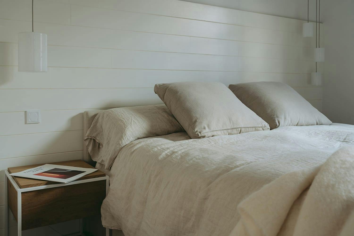The image features a large, well-made bed with a white comforter and pillows. The bed is situated in a room with white walls, creating a clean and minimalist atmosphere. A small table is placed next to the bed, likely for holding a book or other items.

In addition to the bed and table, there are two books on the table, one closer to the left side and the other towards the right side. A remote control is also visible on the table, possibly for controlling the television or other electronic devices in the room.