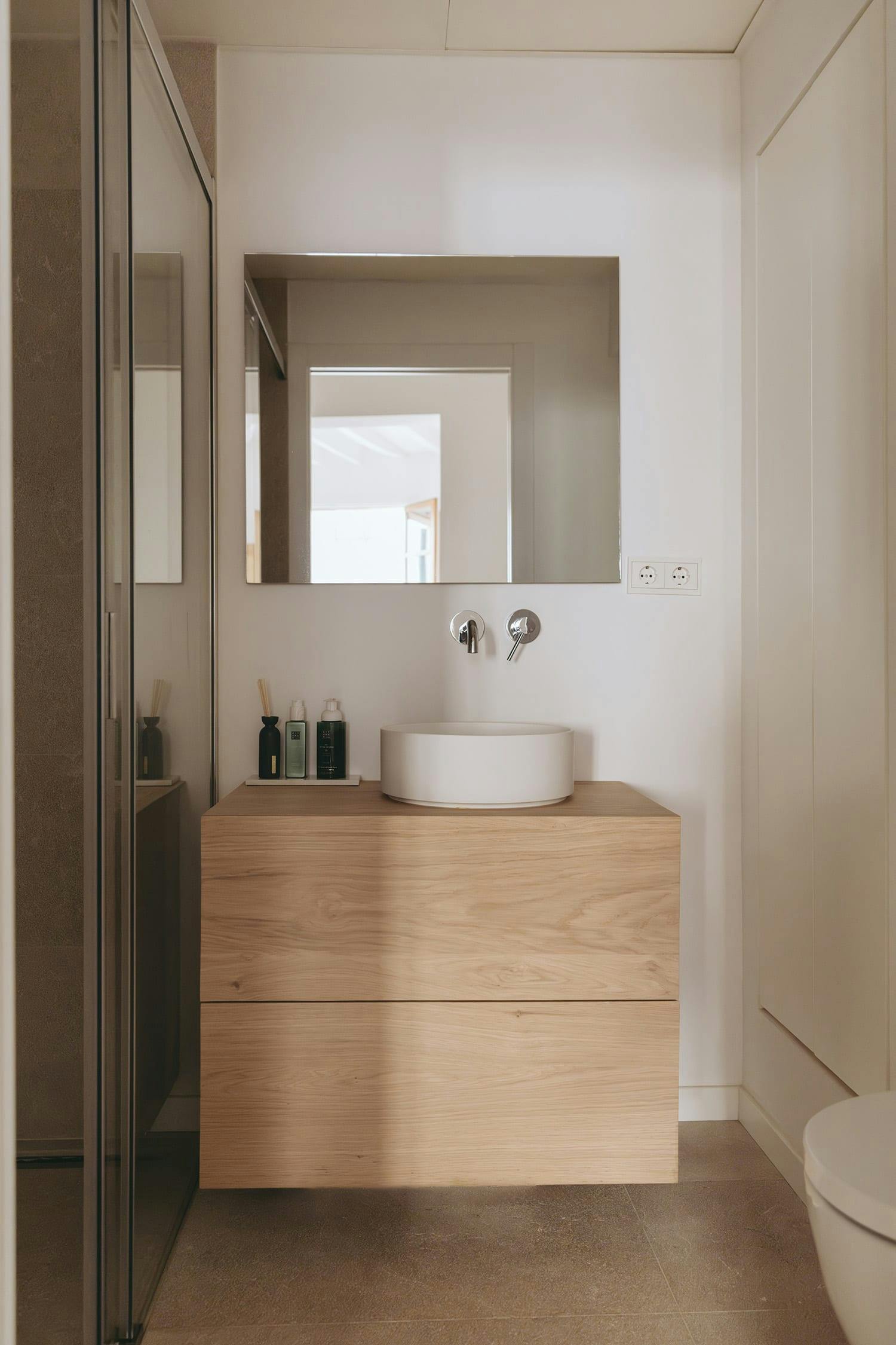 The image features a modern bathroom with a white sink, a mirror, and a wooden vanity. The vanity is made of wood and has a mirror above it. The bathroom is clean and well-organized, with a toilet located in the corner.

In addition to the main sink, there is a small wooden cabinet or shelf above it, which is also white. The bathroom appears to be well-lit, with a window providing natural light. The overall design of the bathroom is minimalistic and clean, making it suitable for a modern and minimalist aesthetic.