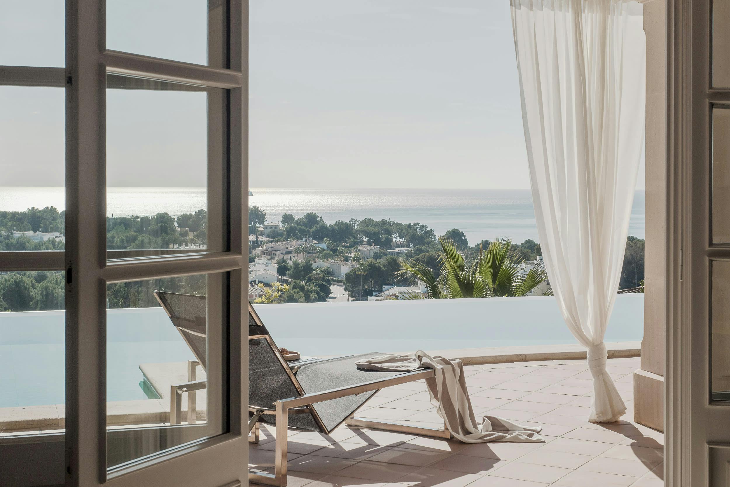 The image features a large, open patio with a view of the ocean. The patio is furnished with a lounge chair and a white lawn chair, both placed near a large window that offers a stunning ocean view. The chair is positioned close to the window, allowing the occupants to enjoy the scenery while relaxing.

In the background, there are several boats visible on the water, adding to the picturesque scene. The patio is surrounded by palm trees, creating a serene and inviting atmosphere.