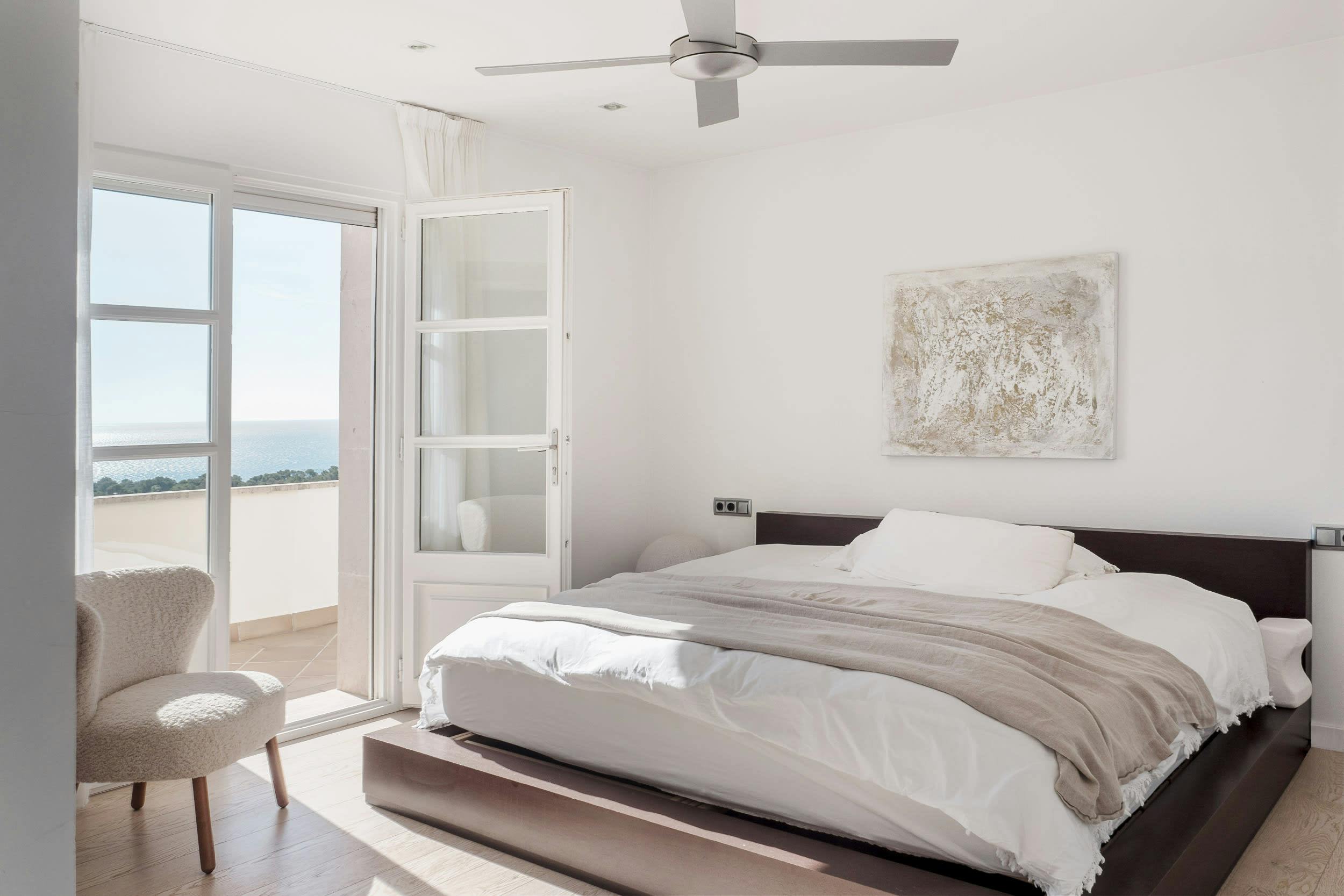 The image features a large, modern bedroom with a white bed and a white headboard. The bed is neatly made, and there is a white comforter on the bed. The room has a large window that offers a view of the ocean, creating a serene and picturesque atmosphere.

A chair is placed near the bed, providing a comfortable seating area for relaxation or reading. A clock can be seen on the wall, adding a touch of functionality to the room. The room also has a ceiling fan, which helps circulate air and maintain a comfortable temperature.

In addition to the b