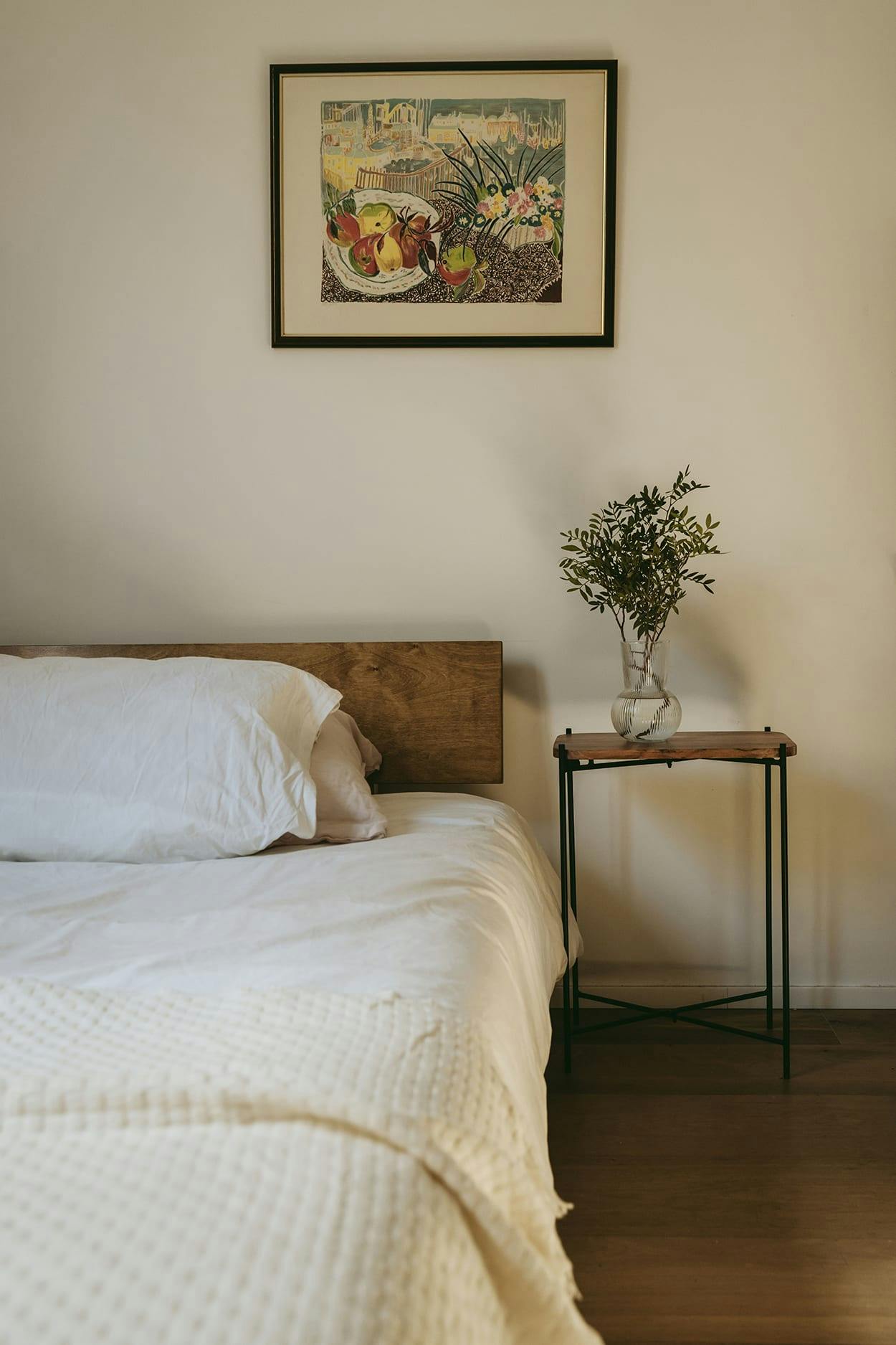 The image features a bedroom with a neatly made bed, a small table with a vase of flowers, and a painting on the wall.