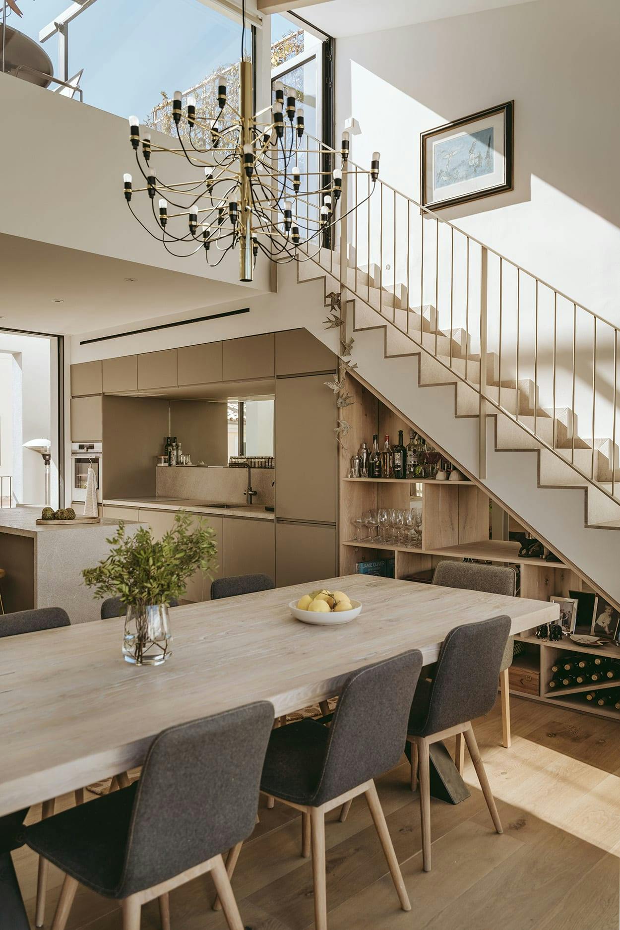 The image features a large, modern kitchen with a wooden dining table and chairs, a chandelier, and a staircase.