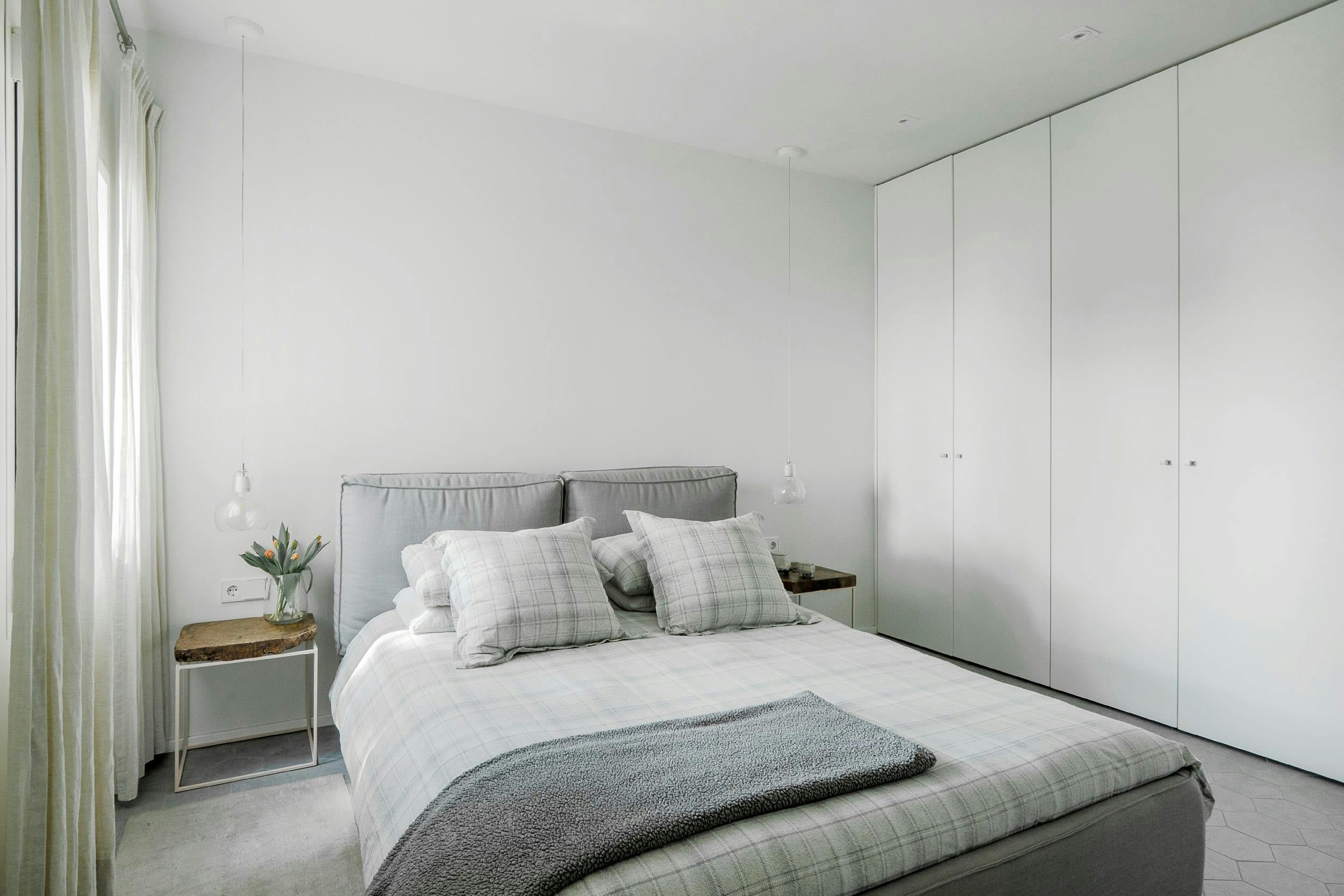 The image features a large bedroom with a neatly made bed, a gray comforter, a white headboard, a white dresser, a white closet, a potted plant, and a vase.