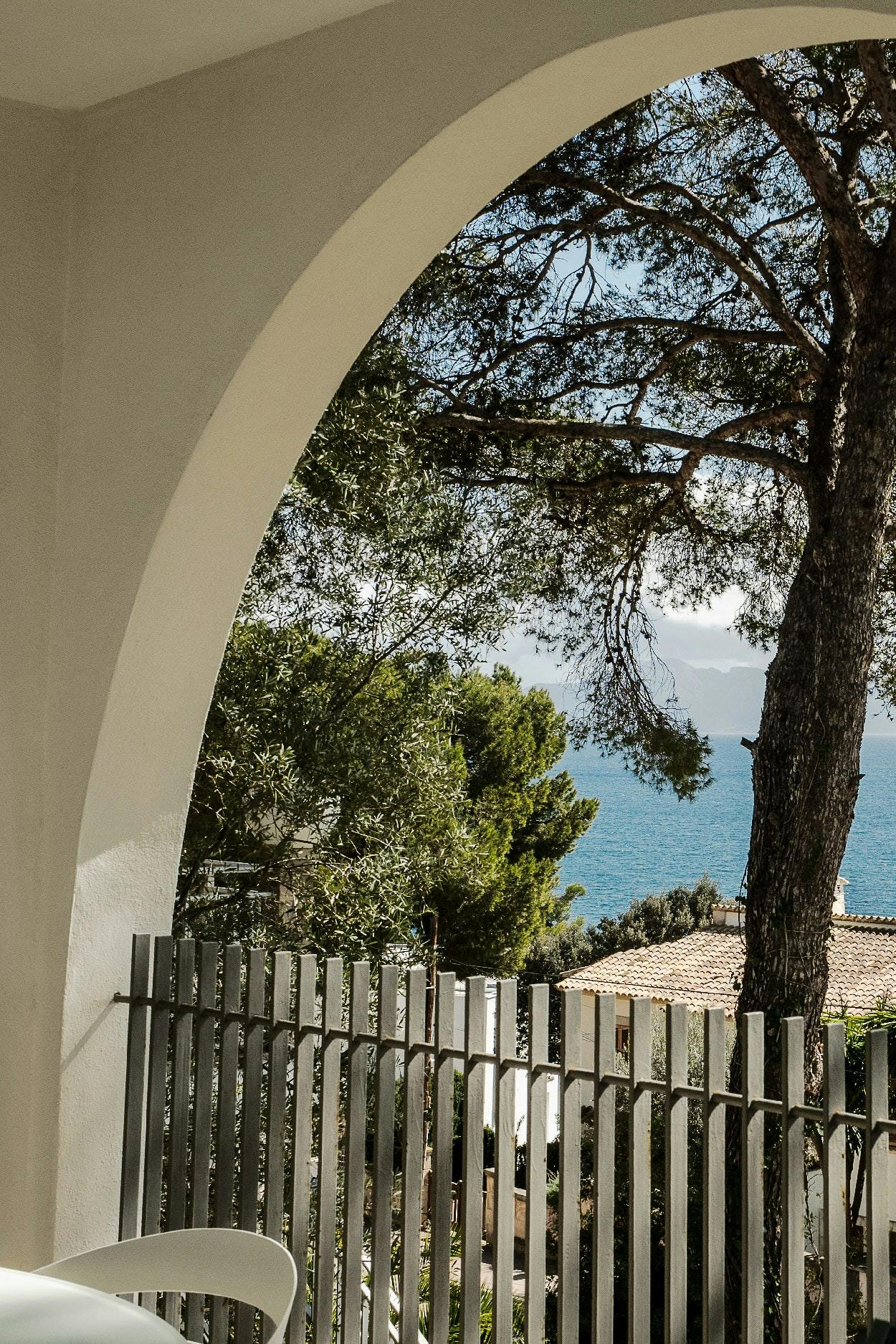 A large tree is in the center of a white fence, with a view of the ocean through a large archway.