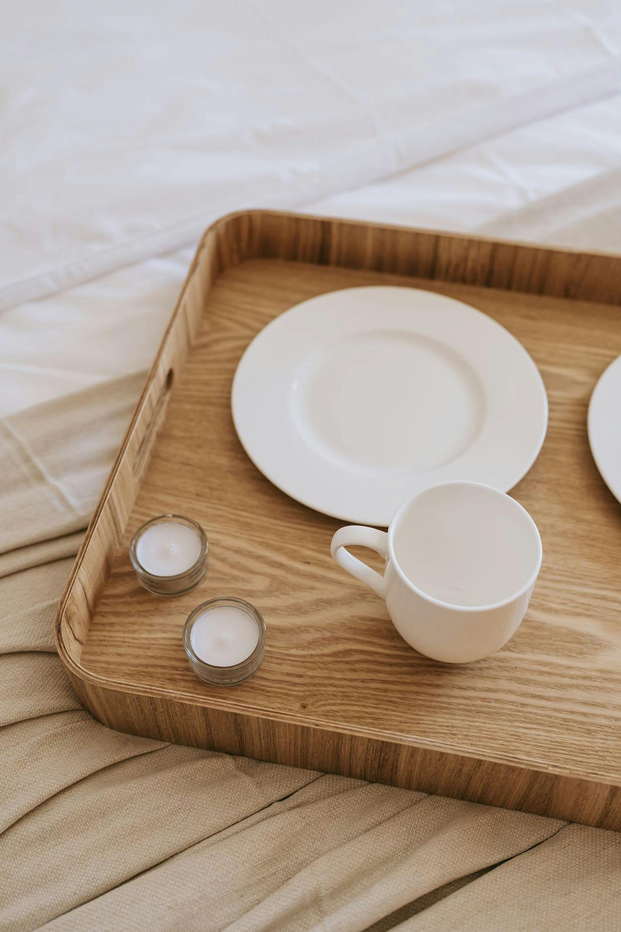 A wooden tray with a white plate, a cup, and a spoon is placed on a bed, creating a simple and elegant dining setup.
