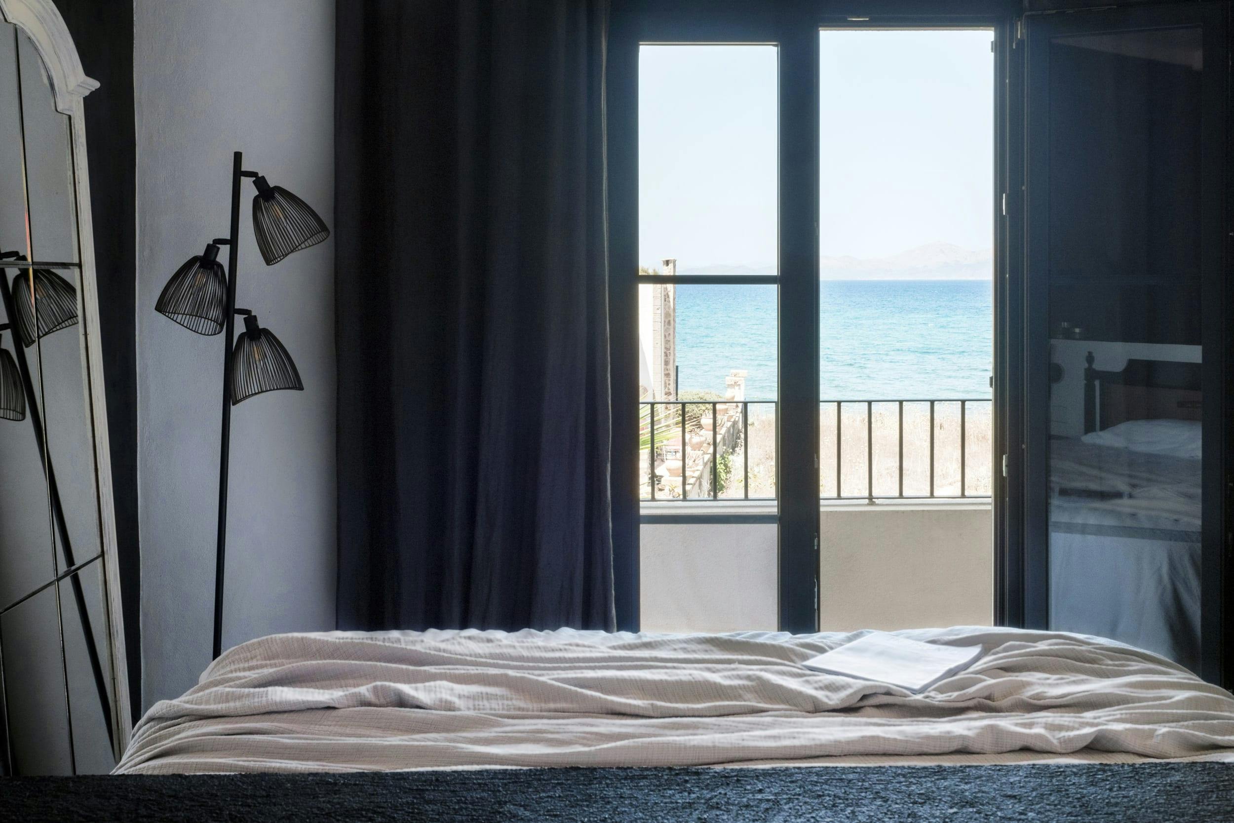 The image features a neatly made bed with a white comforter and a blue blanket, situated next to a large window overlooking the ocean.