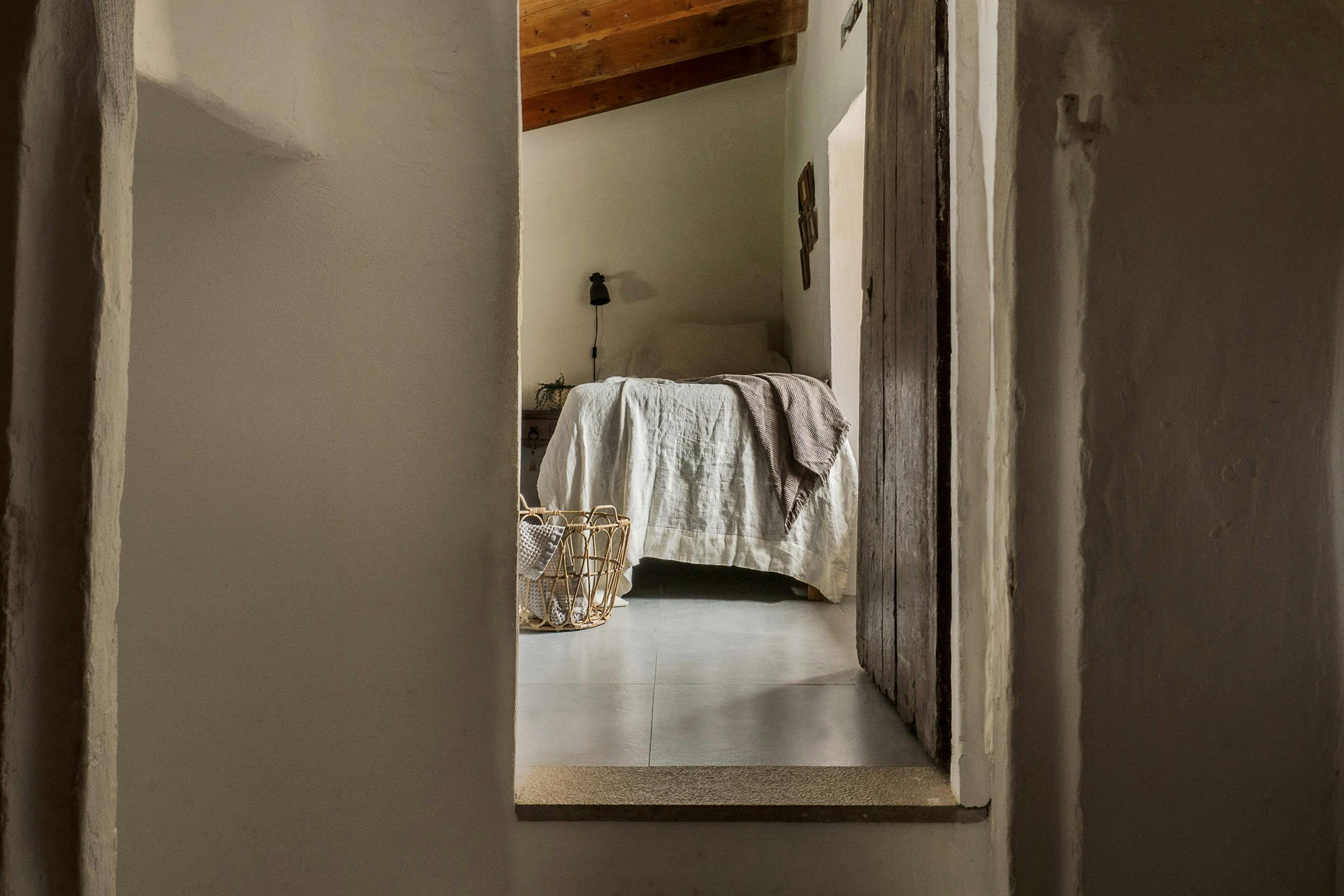 A small, narrow hallway with a mirror on the wall reflects the image of a bedroom with a bed, a towel, and a cup.