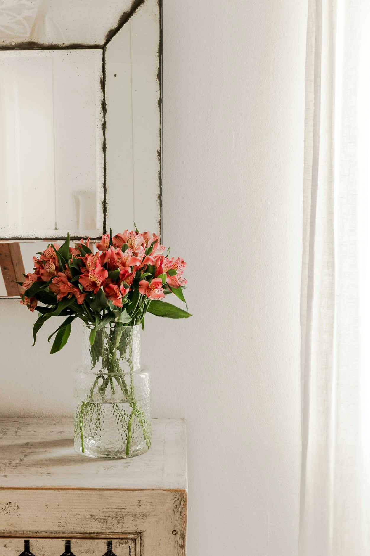 A vase filled with red and orange flowers is placed on a small table next to a mirror, creating a visually appealing and harmonious arrangement.