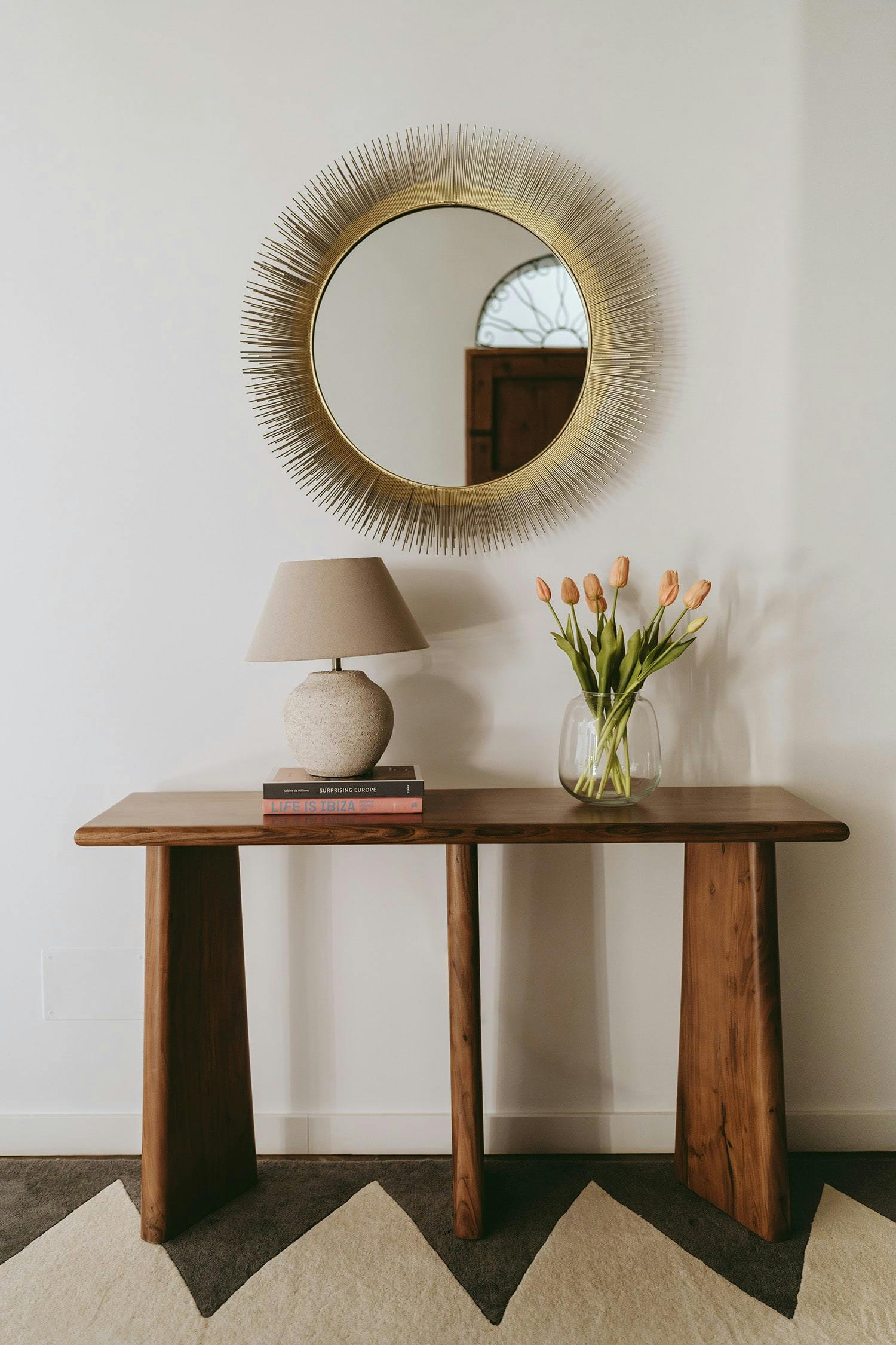 A wooden table with a mirror and a vase of flowers is placed in front of a white wall, creating a visually appealing and harmonious arrangement.