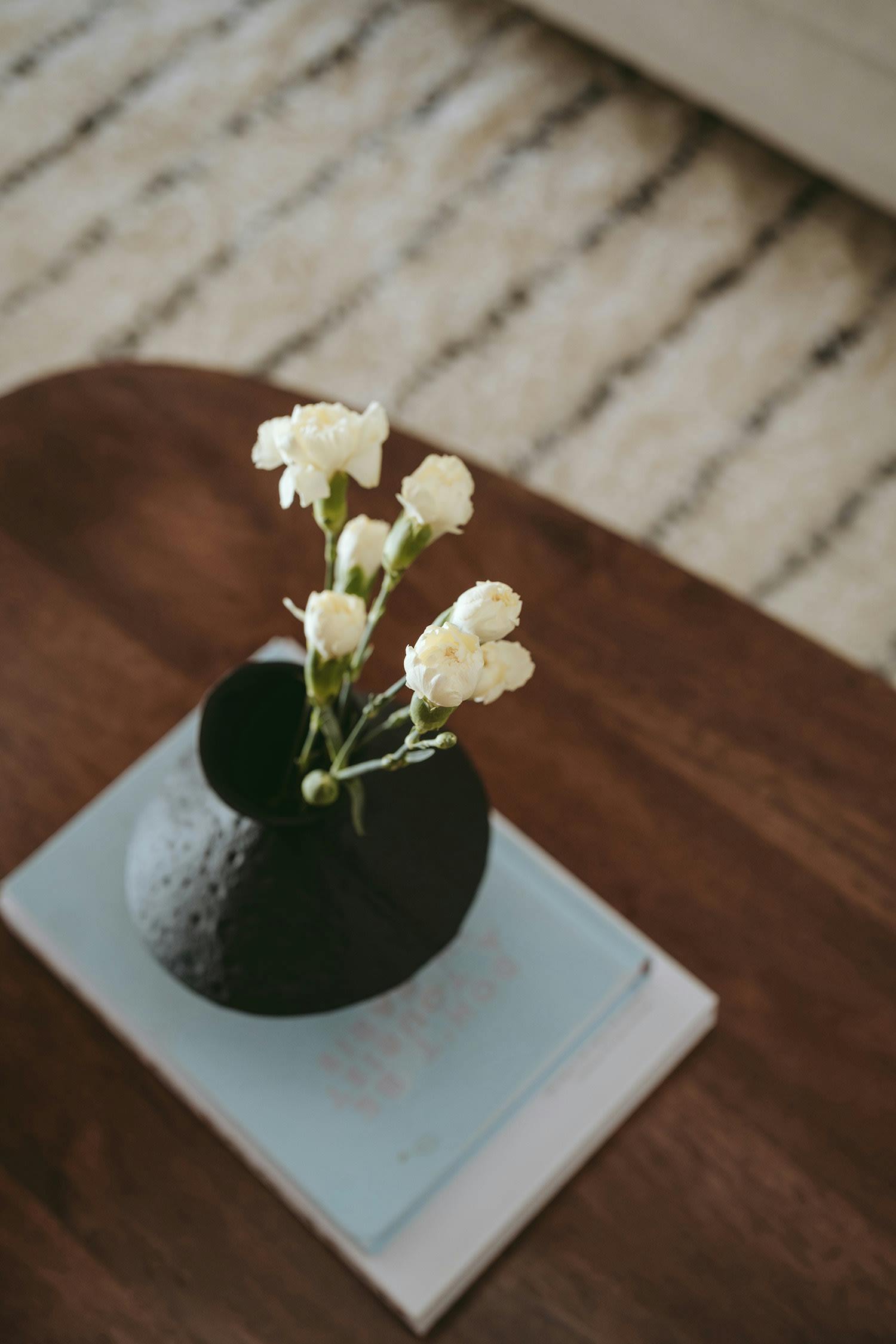 A small vase with white flowers is placed on a table, with a book and a small tablecloth nearby.