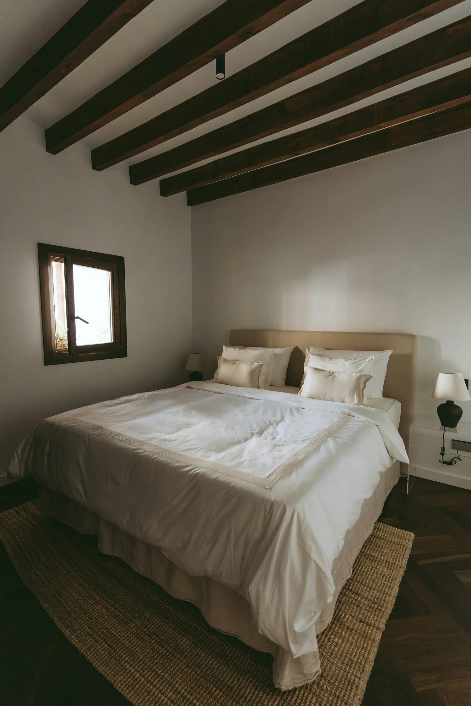The image features a large bedroom with a neatly made bed, a wooden headboard, and a window.