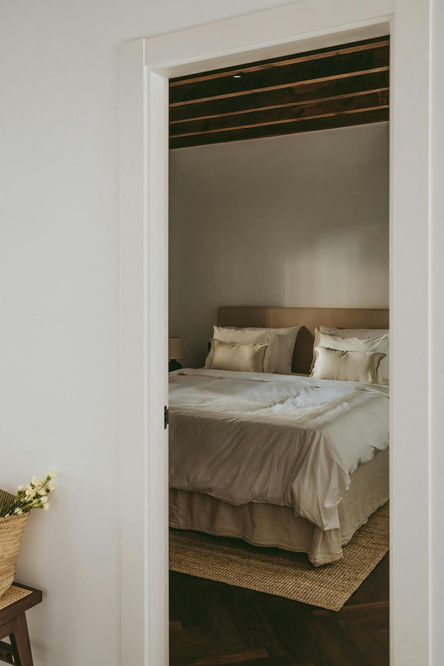 The image features a bedroom with a large bed in the center, surrounded by a white wall and a doorway. The bed is neatly made with a white comforter and pillows, and there is a potted plant nearby.