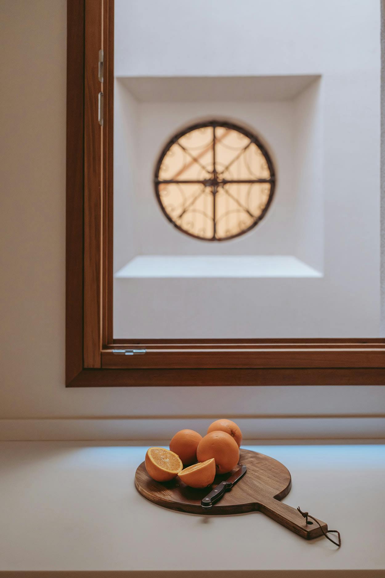 A window with a large, round, stained glass window is open, revealing a table with a bowl of oranges and a knife on it.