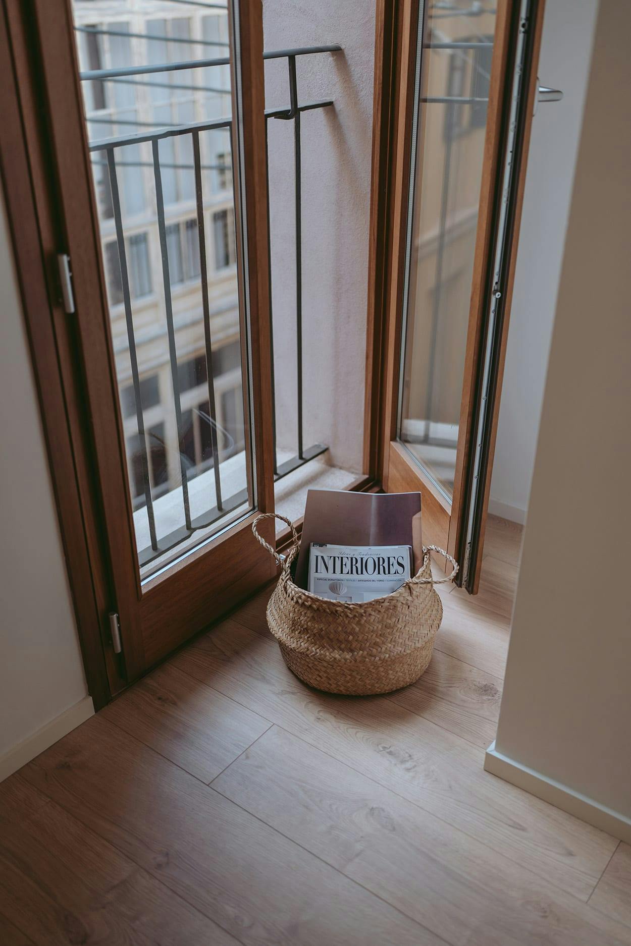 A small basket is placed on a wooden floor next to a window, with a cat sitting inside.