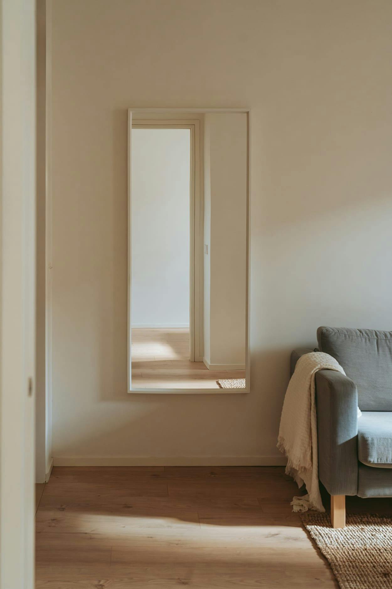 A small, clean, and empty room with a couch and a chair in the corner, a window with a view of the outside, and a doorway leading to another room.