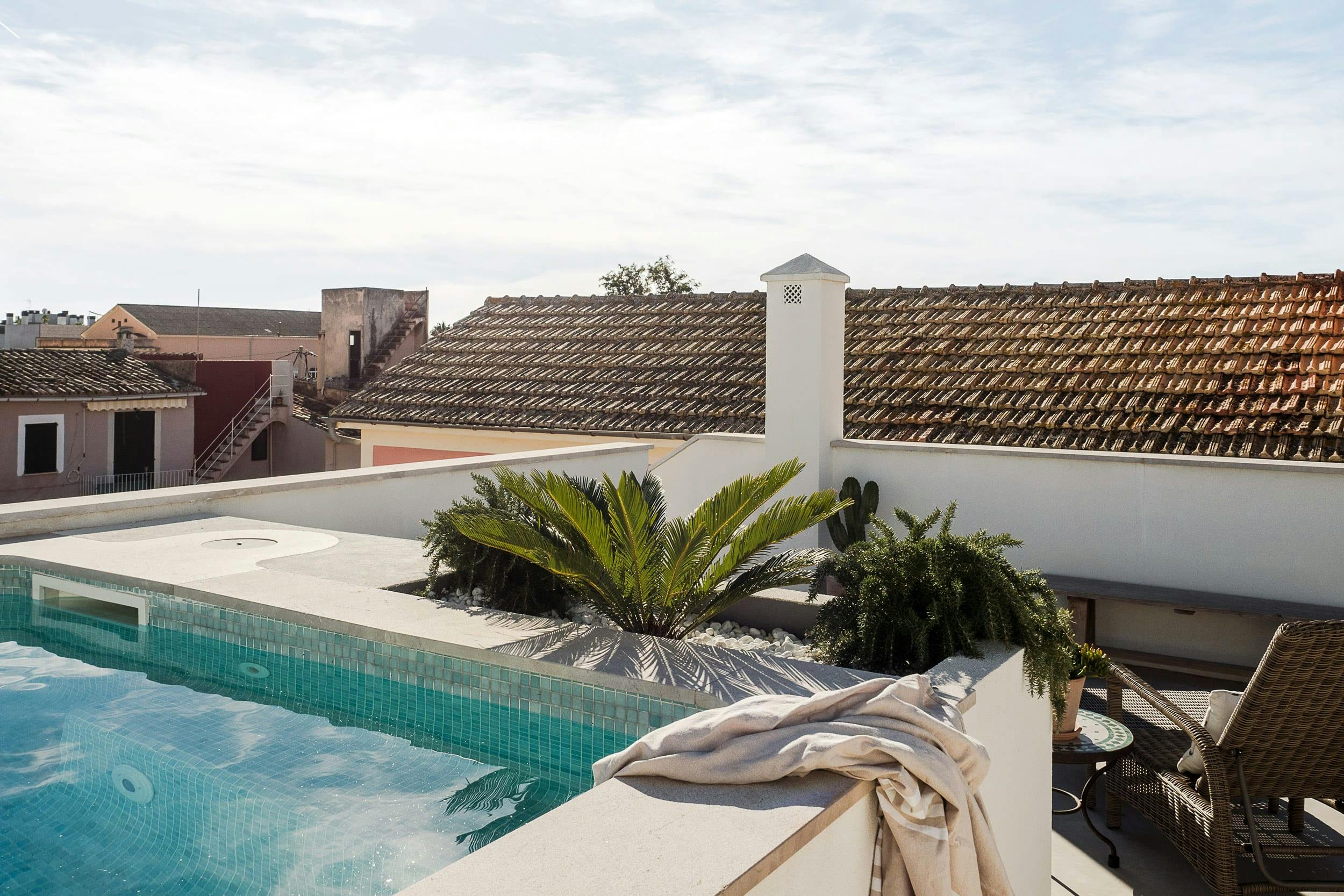The image features a large swimming pool with a white tile roof, surrounded by a patio area with a lounge chair and a table.