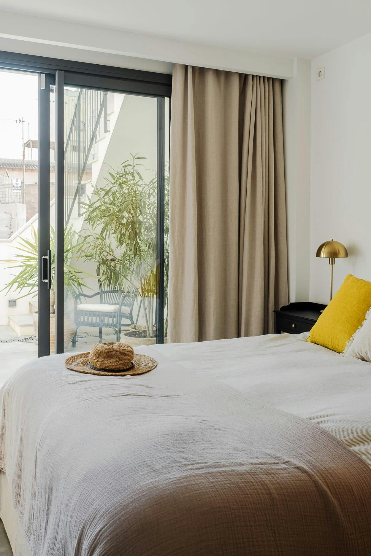 The image features a neatly made bed with a white comforter and a yellow pillow, placed next to a window with a view of a city. There is a chair and a potted plant nearby, and a handbag is also present in the room.