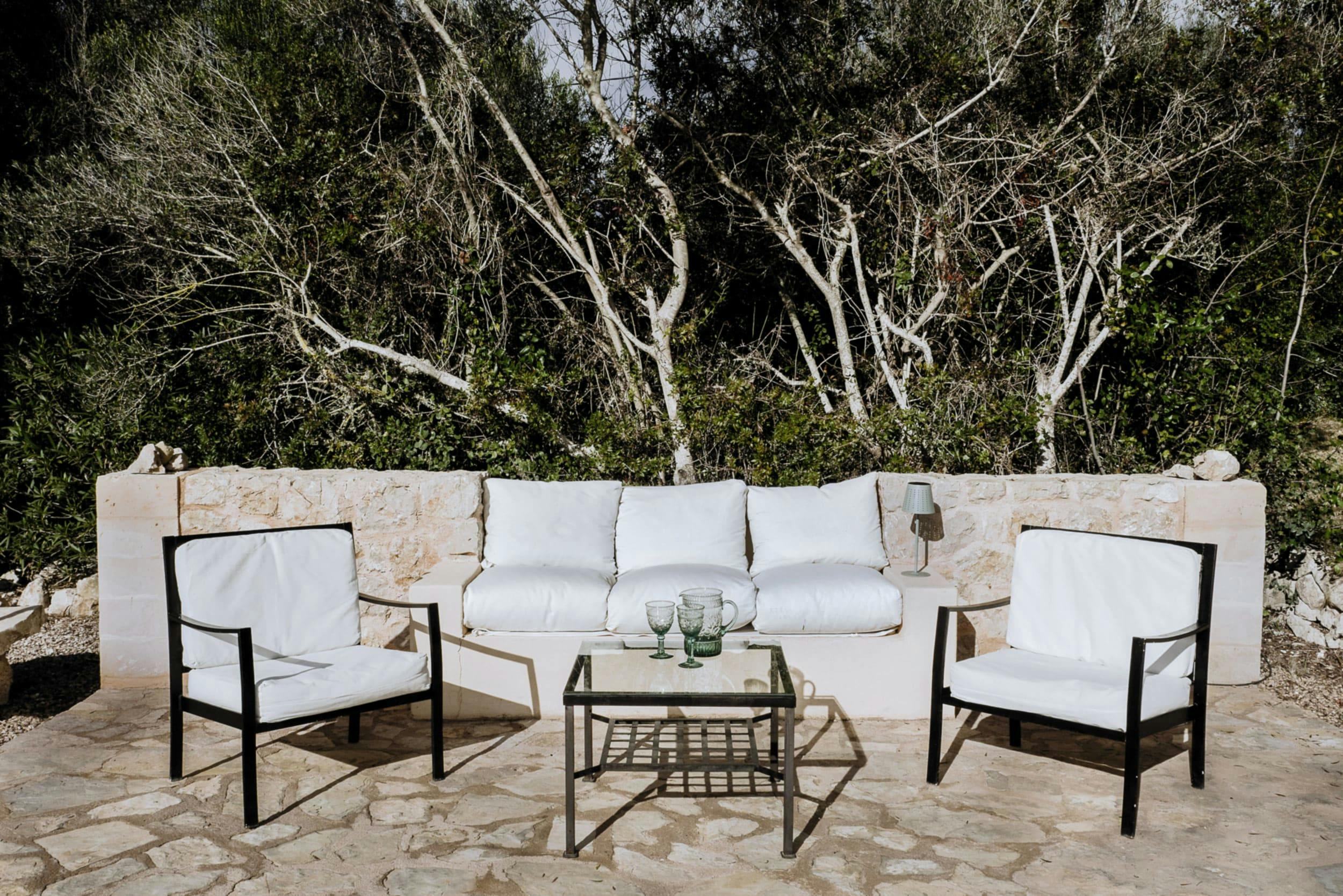 The image features a patio with two white chairs and a table, creating a cozy outdoor seating area. The chairs are placed close to each other, and the table is situated between them. There are also two potted plants, one on the left side and another on the right side of the table, adding a touch of greenery to the scene.