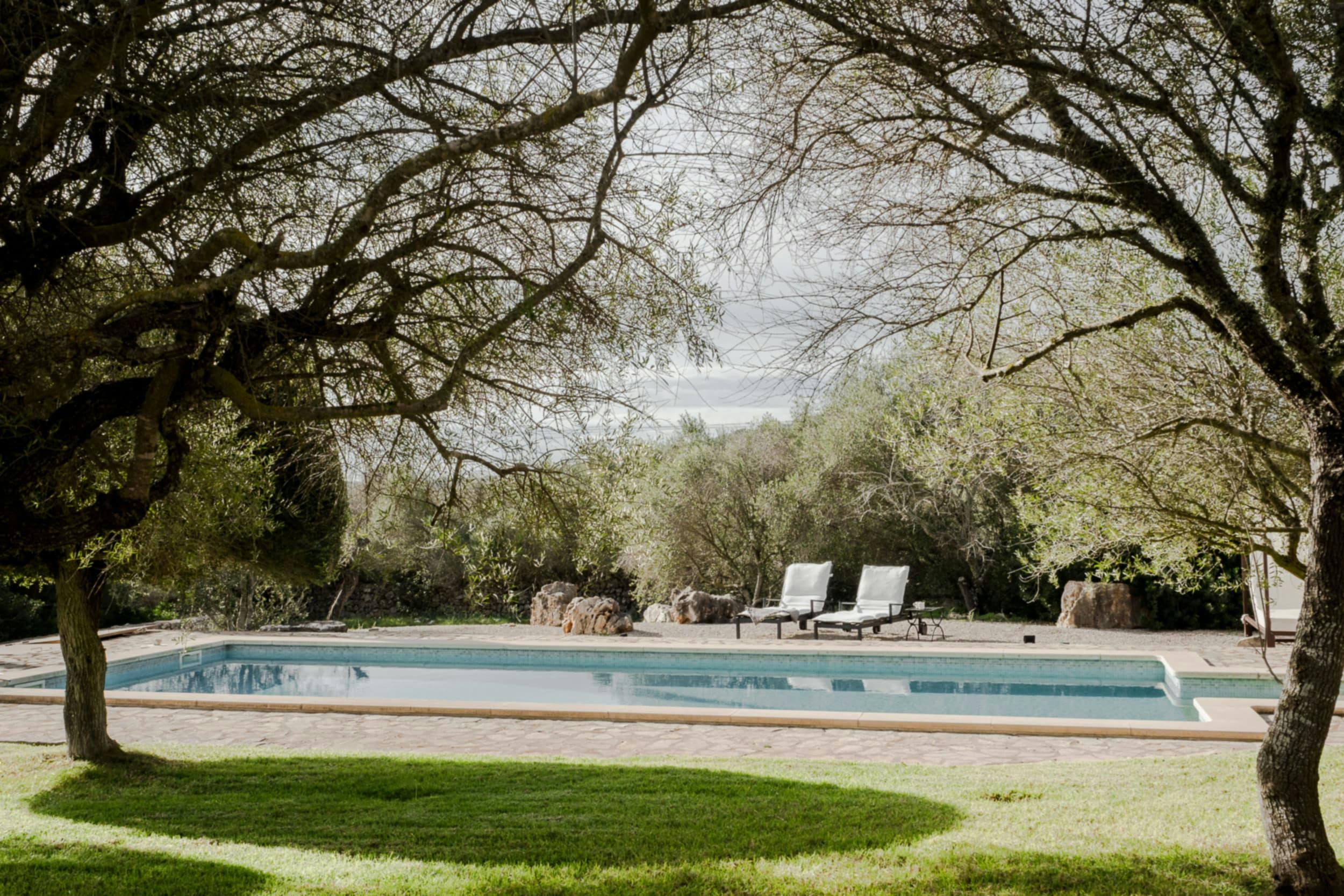A large swimming pool is surrounded by a lush green lawn, with a patio area and chairs nearby.