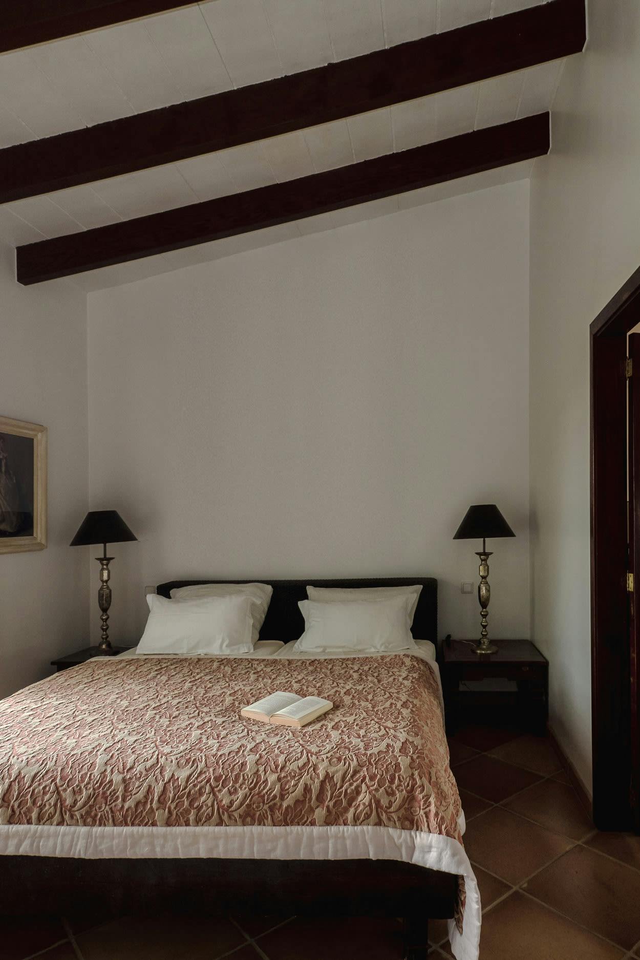 The image shows a bedroom with a neatly made bed, two lamps, a nightstand, and a chair.