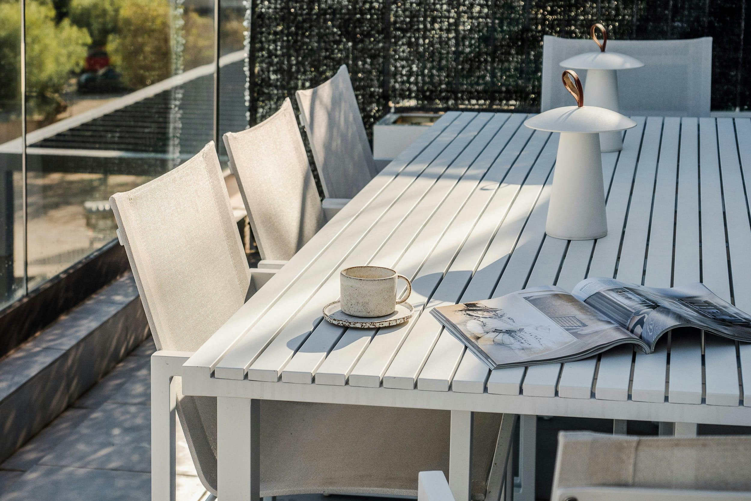 A white table with a cup of coffee, a book, and a napkin is set up on a patio, with a chair nearby.