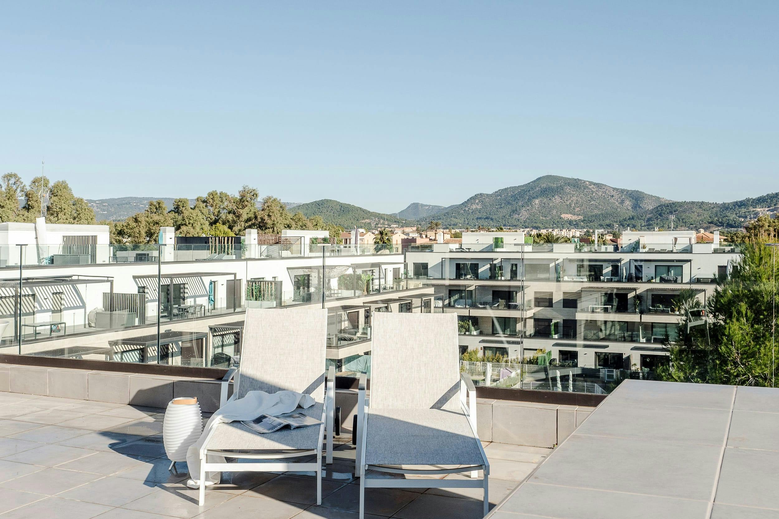 The image features a large, modern apartment building with a balcony on the second floor, overlooking a cityscape with mountains in the background. The balcony is furnished with a white chair, a couch, and a dining table, creating a comfortable outdoor seating area.