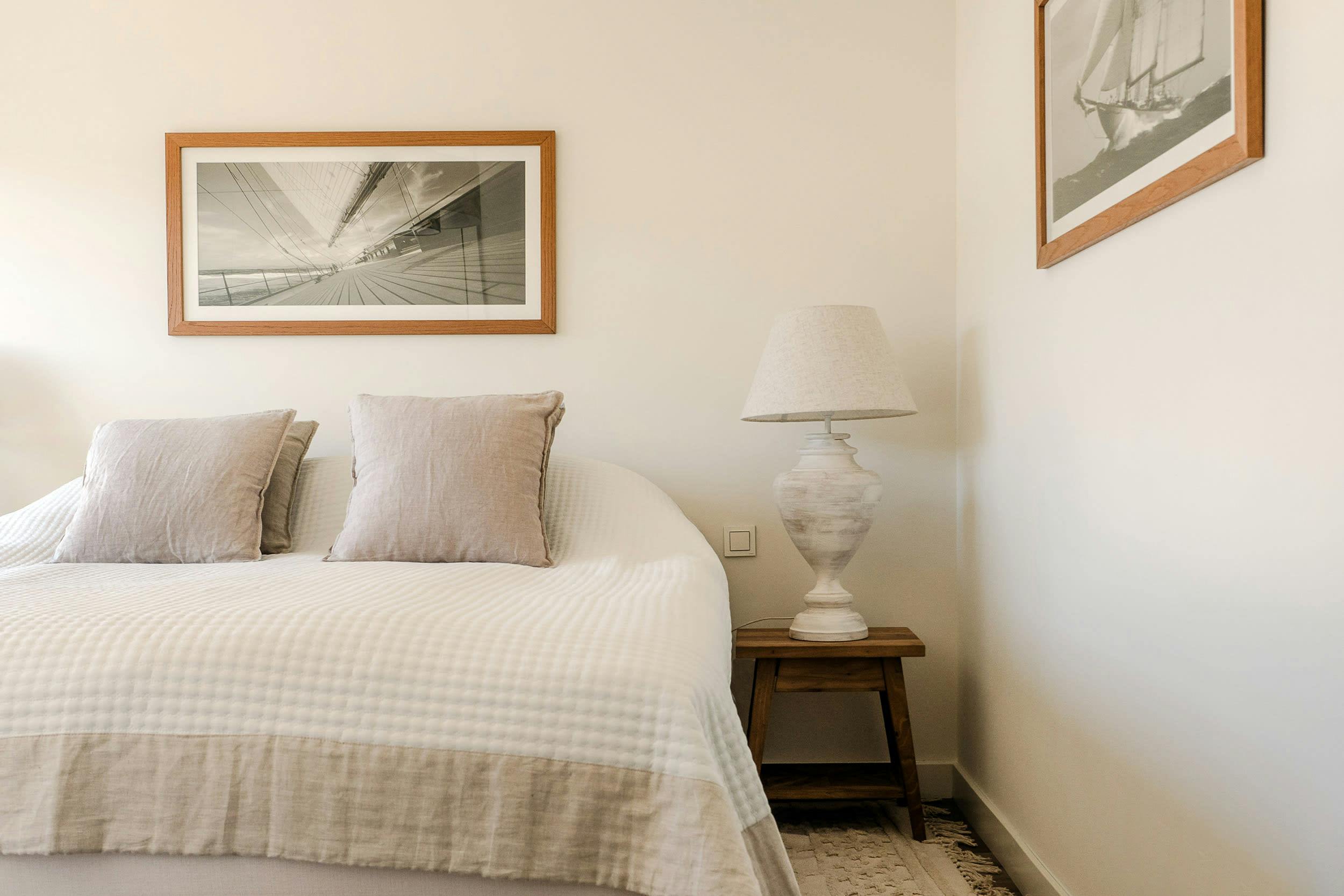 The image features a neatly made bed with a white comforter and pillows, placed next to a lamp and a picture on the wall.