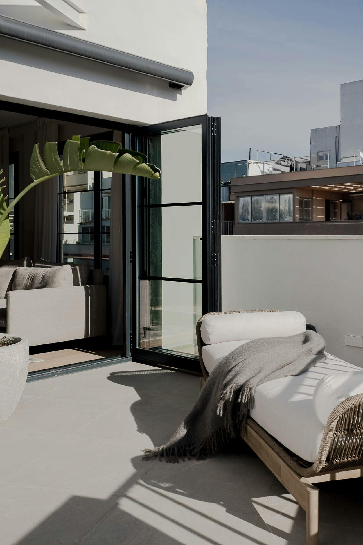 The image features a large, modern, and well-lit room with a white color scheme, featuring a large bed, a couch, and a chair. The room is located on a rooftop with a view of the city, and there are potted plants and a vase in the room.