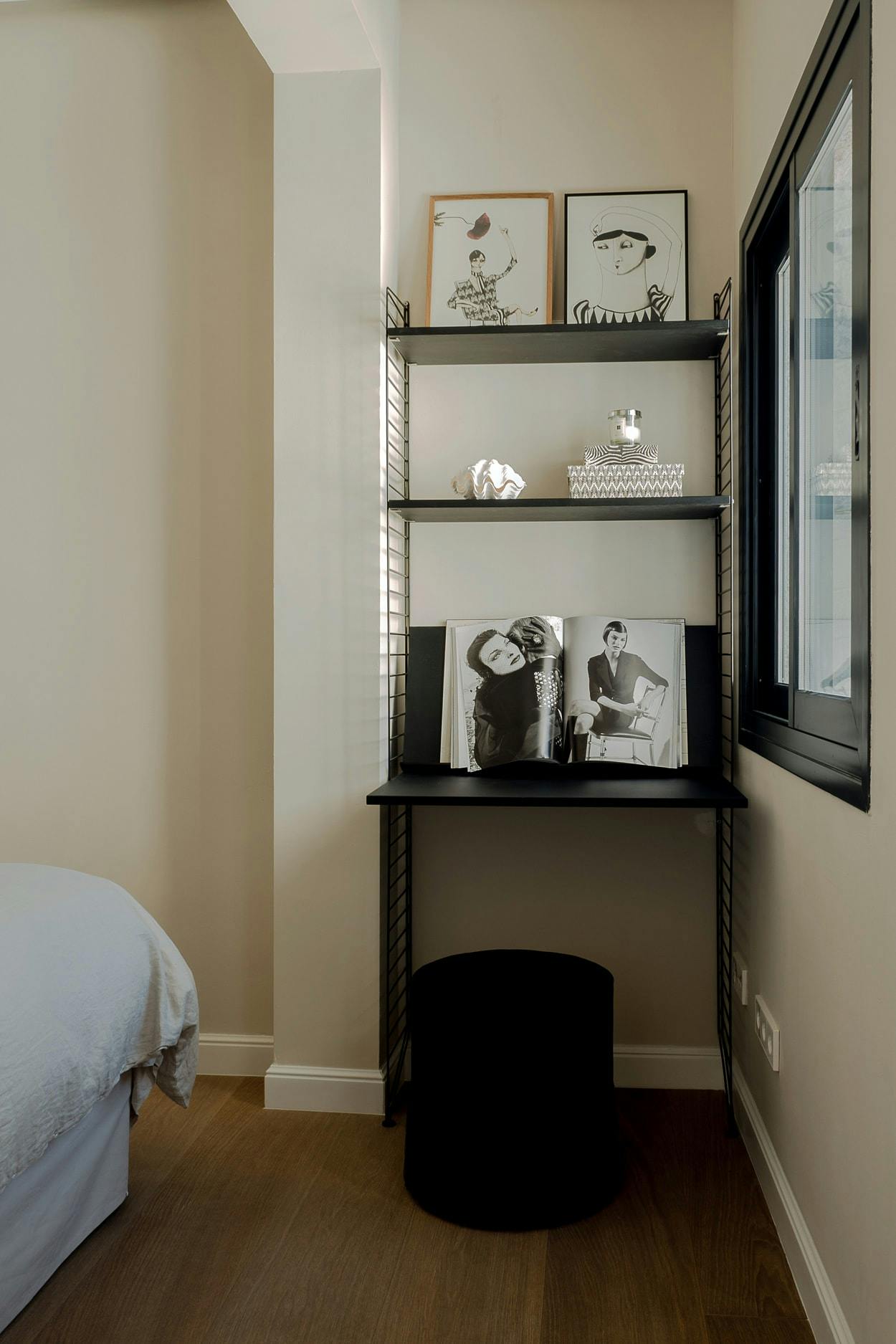 The image features a bedroom with a bed in the corner, a black bookshelf with a black book on top, and a window.