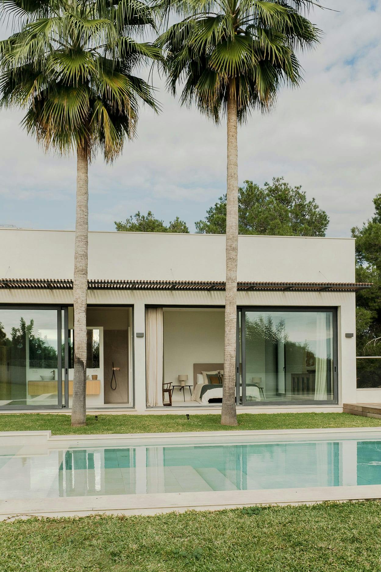 A large, modern house with a swimming pool is surrounded by palm trees, creating a tropical atmosphere. The house has a large glass door and a white exterior, and there are several chairs and a dining table placed around the pool area.