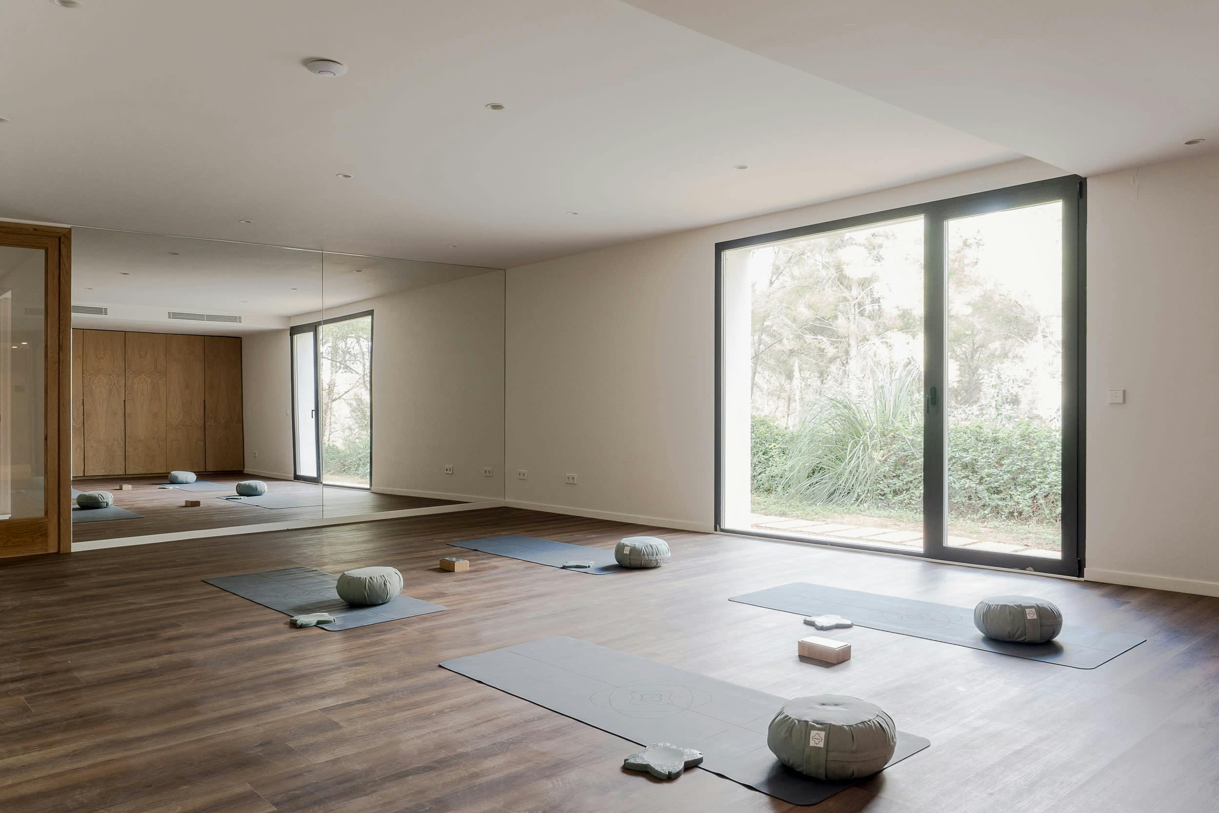 A large room with a wooden floor and a large window is filled with a variety of yoga mats and balls, creating a spacious and inviting atmosphere for practicing yoga.
