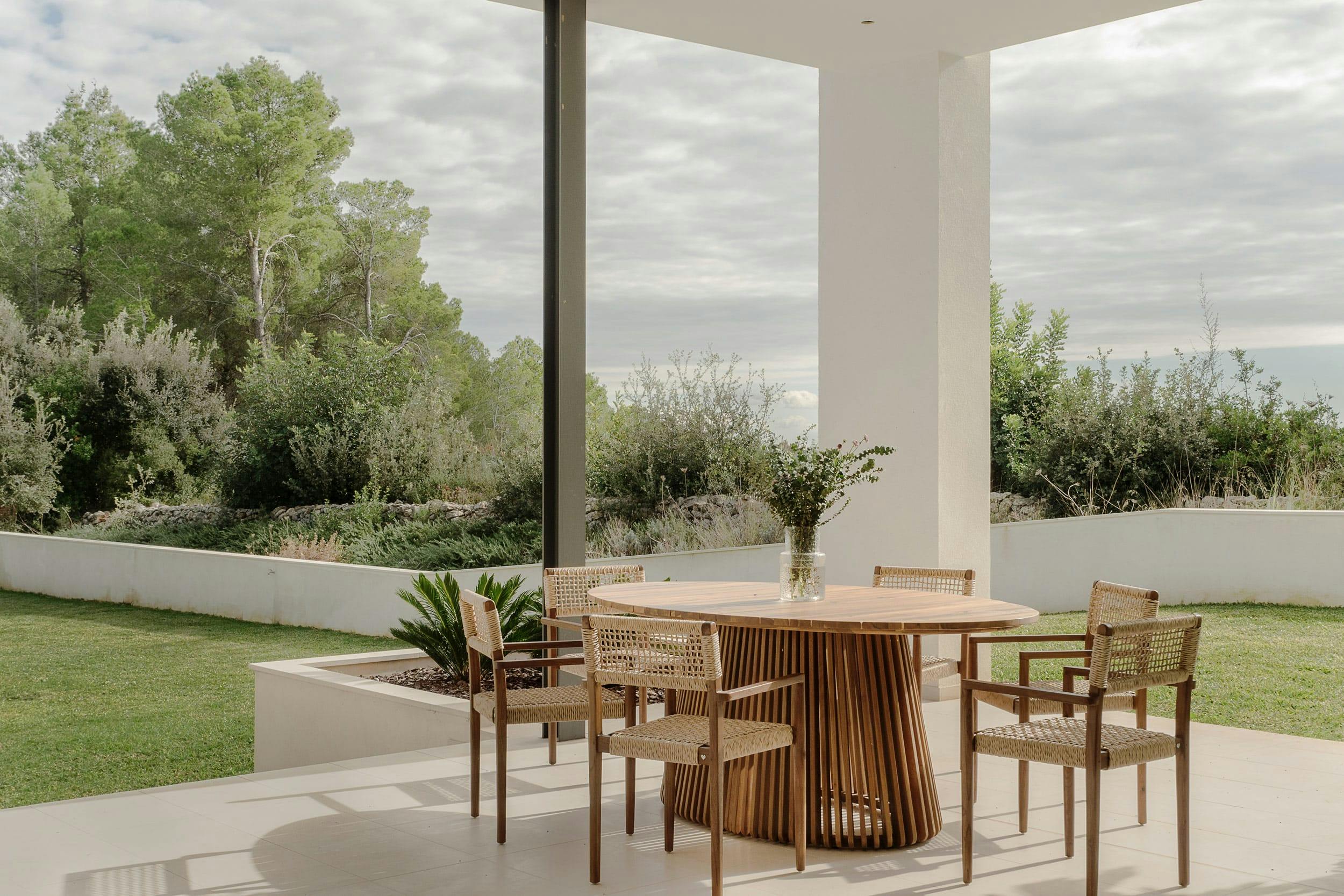 The image features a large glass window with a view of a patio area, which is furnished with a wooden dining table and chairs. The table is surrounded by four chairs, and there are two potted plants placed on the table and the patio.