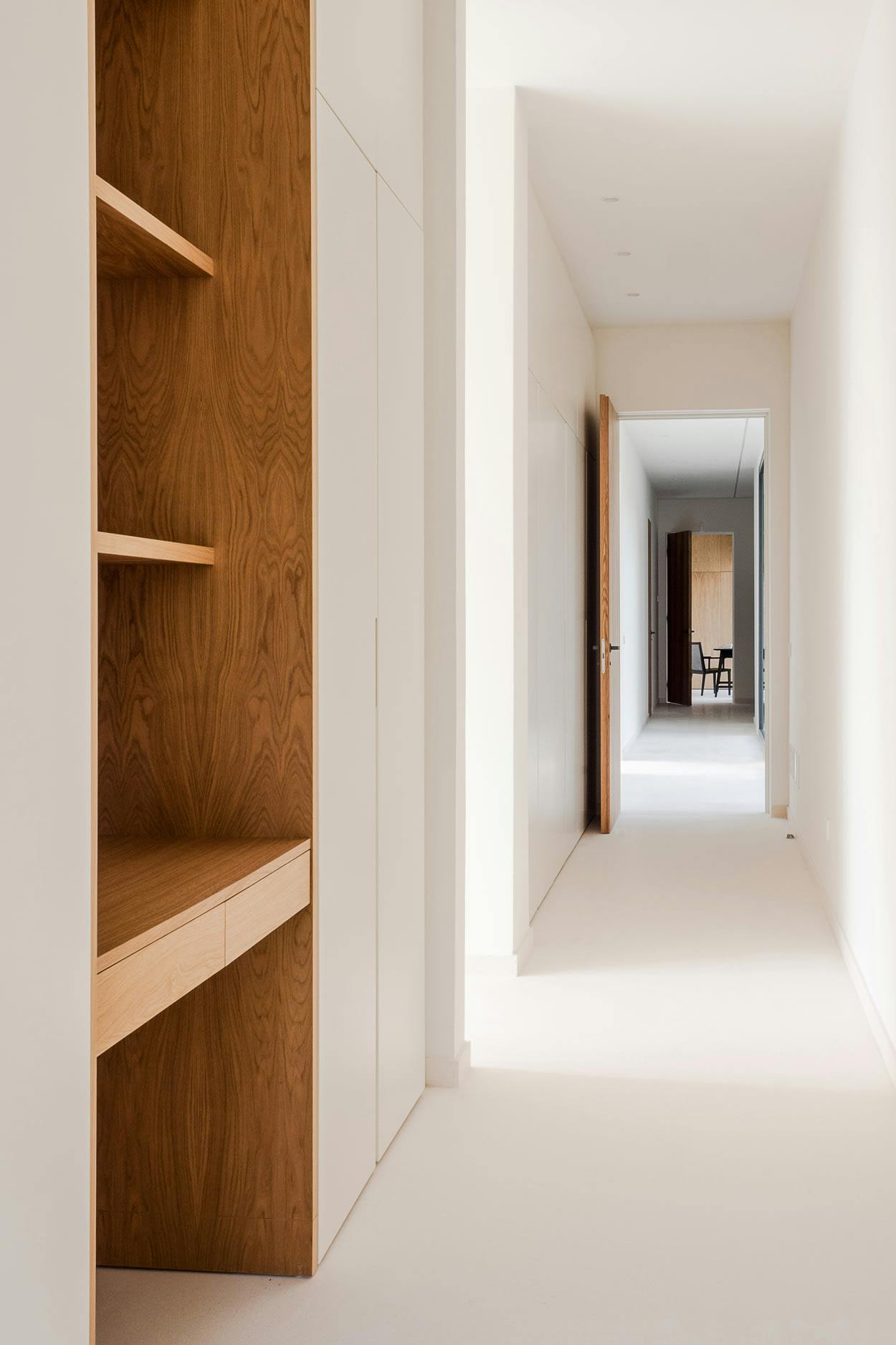 A long, narrow hallway with white walls and wooden doors is shown in the image.